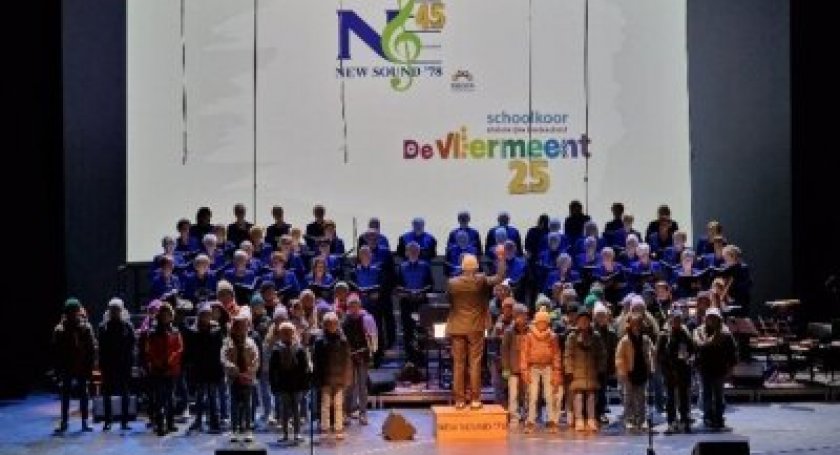 Op de afbeelding zie je het schoolkoor de Vliermeent en Zoetemeers gemengd koor 'New Sound '78'. Ze staan samen op een groot podium met in het midden dirigent Kees van Venetie.